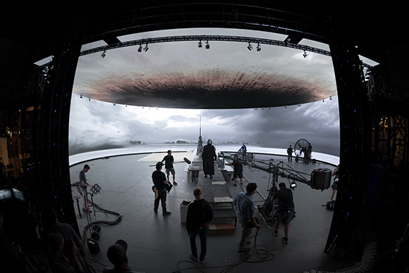 Film crew on a large circular LED stage displaying a cloudy sky and landscape.