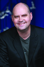 A bald man wearing a black blazer and dark t-shirt smiles against a blue background.