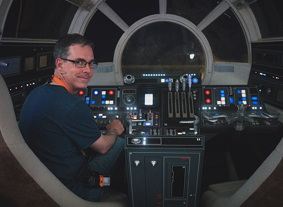 A man with glasses sits in a dark cockpit filled with glowing buttons, levers, and circular windows.