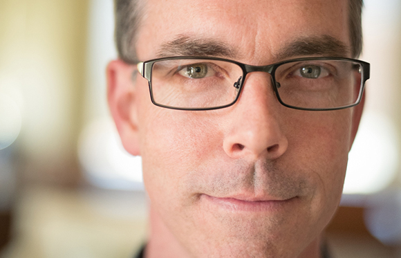 Close-up of a man with short hair and black-rimmed glasses looking directly at the camera.