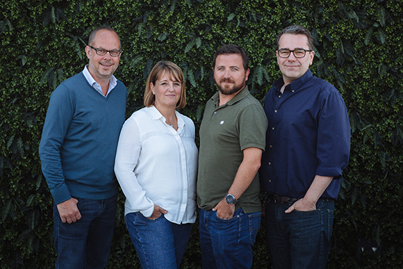 Four adults standing in a row against a dense green leafy wall.