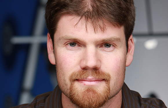Close-up of a man with light eyes and a reddish-brown beard and mustache.