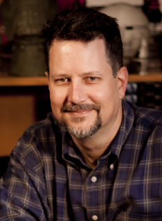 A man with dark hair and a goatee smiling in a blue plaid shirt.