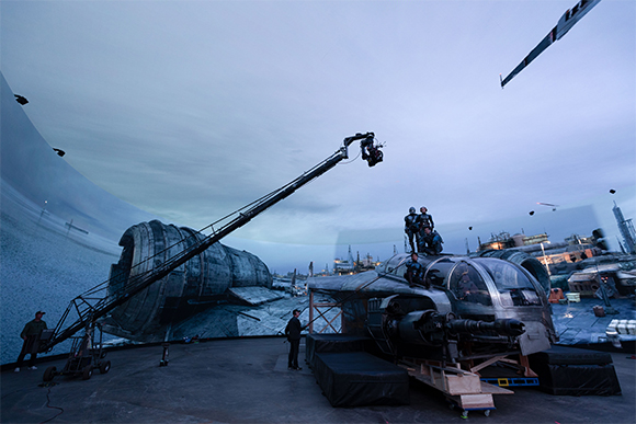 Film crew using a crane camera to film a large spacecraft model in front of a giant curved projection screen.