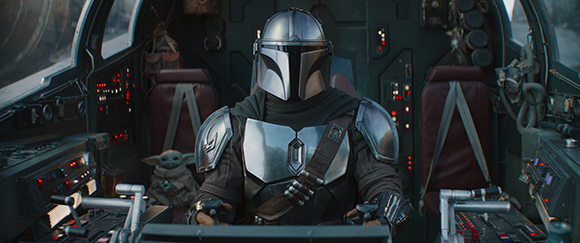 A character in silver Mandalorian armor sits in a spaceship cockpit next to a small green creature.