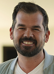 A smiling man with dark hair and a salt-and-pepper beard wearing a light-colored jacket.
