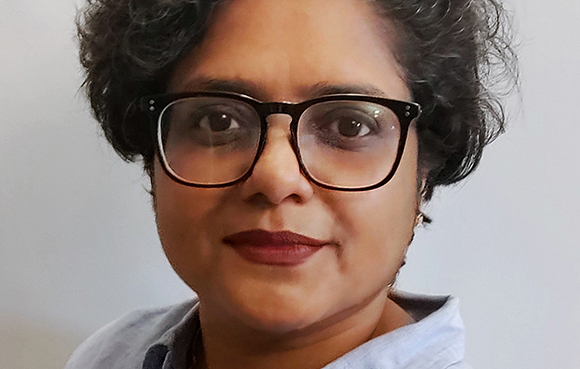 A woman with dark curly hair and black-rimmed glasses looking toward the camera.
