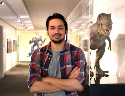 A man with dark hair and a beard stands with arms crossed in a gallery next to a large dinosaur model.