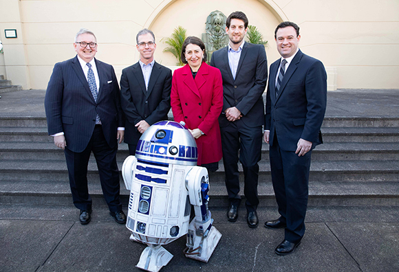 Five people in professional attire stand behind an R2-D2 droid on outdoor stone steps.