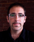 A man with short dark hair, glasses, and a mustache looking forward against a brick wall.