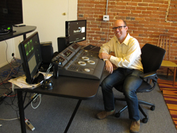 A man in a light shirt and glasses sits at a desk with multiple monitors and a large video editing console.