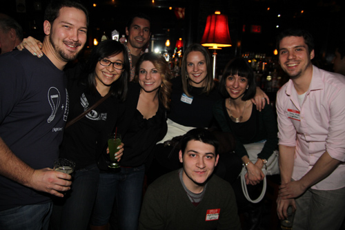 A group of smiling young adults posing together in a dark bar setting.