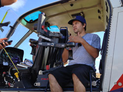 A man in a blue cap and grey shirt sits inside a helicopter cockpit surrounded by controls.