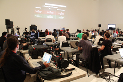 People sit at tables with laptops watching a presentation on a large screen in a lecture hall.