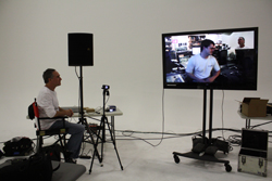 A man in a white shirt sits in a chair watching a large television screen in a studio setting.