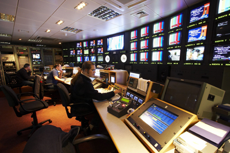 Three technicians sit at desks in a control room surrounded by walls of video monitors.