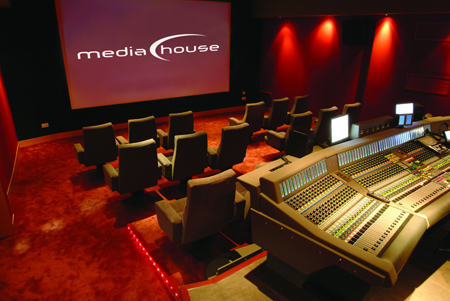 A dark media studio featuring a large projection screen, red walls, and a large audio mixing console.