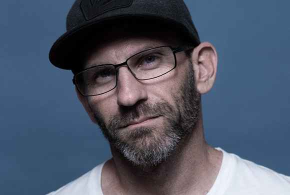A man with a salt-and-pepper beard, wearing a black baseball cap and glasses against a blue background.