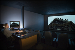 A person sits at a desk with monitors in a dark room facing a large projection screen.