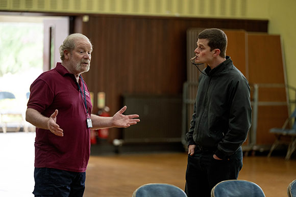A man in a maroon polo shirt gestures with both hands while speaking to a man in a black jacket.