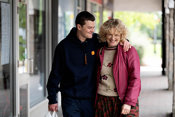 A man in a dark hoodie and a woman in a pink jacket walk side-by-side, smiling.