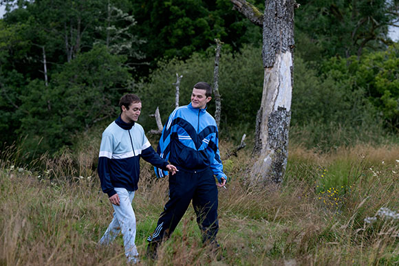 Two men in athletic tracksuits walking through a tall grass field near a forest.