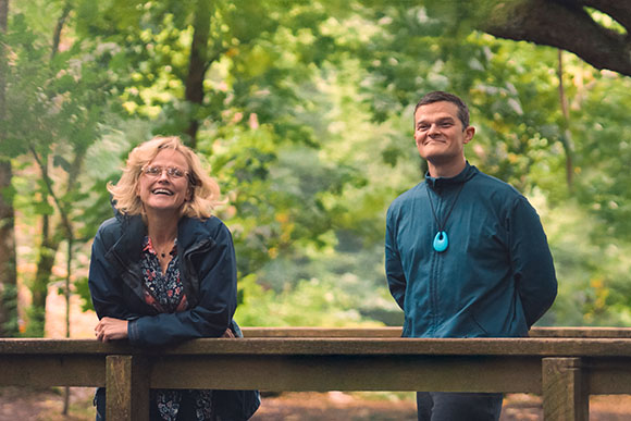 A blonde woman in glasses laughs while leaning on a wooden railing, next to a man in a blue jacket smiling.