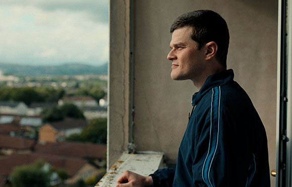A man in a dark blue jacket looks out from a high balcony over a residential area and distant hills.