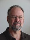 A man with glasses and a grey-flecked goatee looking toward the camera against a plain white background.