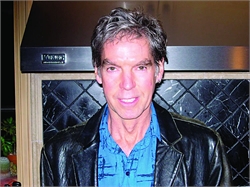A man with salt-and-pepper hair wearing a black leather jacket over a blue patterned shirt.