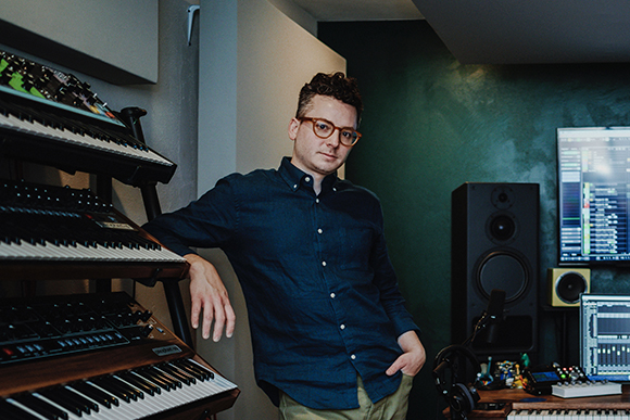 A man with glasses in a blue shirt leans against a stack of keyboards in a music studio.