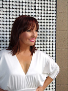 A woman with reddish-brown hair and a white blouse smiling against a silver sequin background.