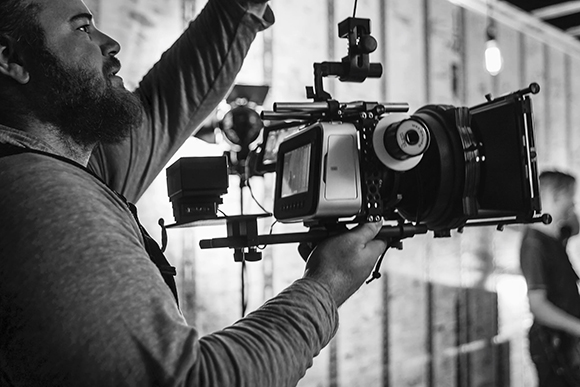 A bearded man holds and operates a large professional cinema camera with a monitor and matte box.