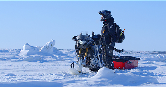 A person in winter gear stands beside a snowmobile and a red sled on a vast, snow-covered plain under a blue sky.