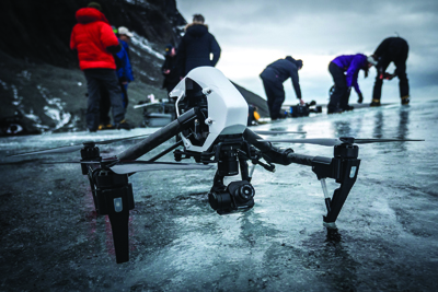 A black drone rests on a frozen, textured surface with blurred figures standing in the distance.