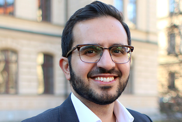 A man with dark hair, glasses, and a beard smiling against a blurred building background.