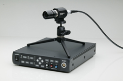 A black electronic base unit with a small camera mounted on a tripod stand and a connected black cable.