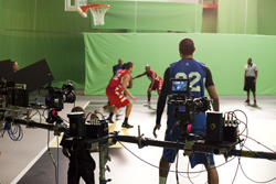 A film crew with cameras and equipment filming basketball players practicing in an indoor gym.