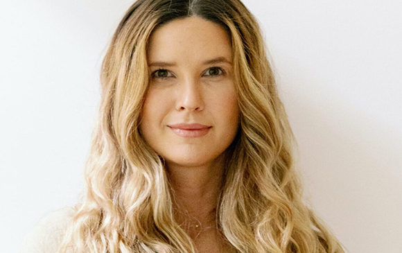 A woman with long, wavy blonde hair looks directly at the camera against a plain white background.