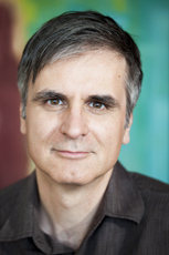 A man with short dark hair and brown eyes looking forward against a blurred green and red background.