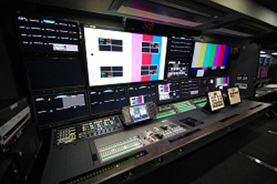 A broadcast control room featuring a large wall of monitors displaying color bars and technical data above a control desk.