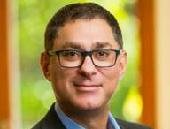A man with short dark hair and glasses smiling against a blurred green background.
