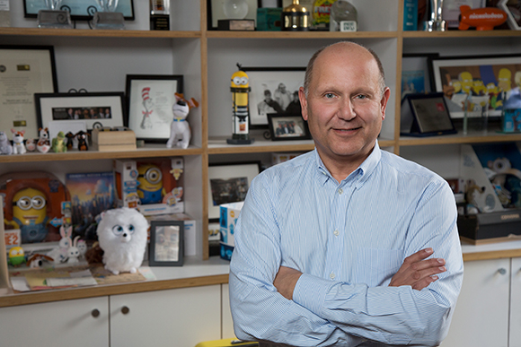 A man in a light blue button-down shirt stands with arms crossed in front of shelves of toys and awards.