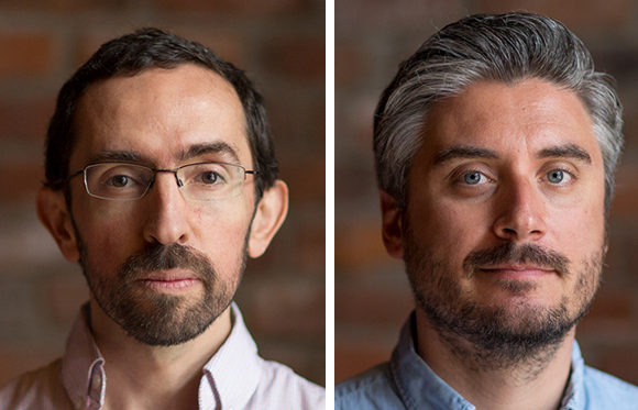 Two men with facial hair looking directly at the camera against a brick background.