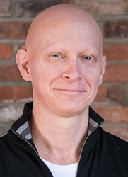 A bald man with blue eyes smiling against a brick wall background.