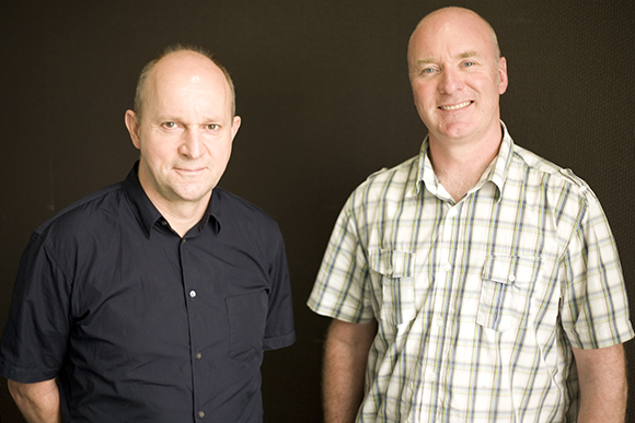 Two men, one in a dark blue shirt and one in a plaid shirt, smiling against a dark background.