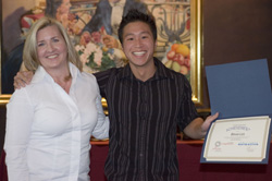A blonde woman in a white shirt and a man in a striped shirt smiling and holding an open blue award folder.