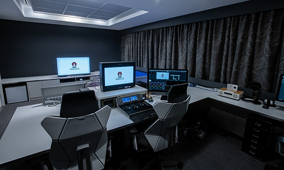 A dark media studio featuring two workstations with monitors, control panels, and ergonomic chairs.