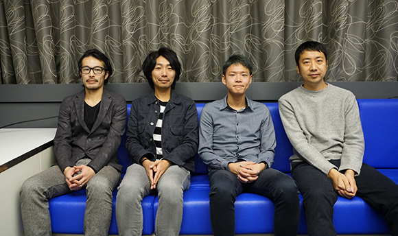 Four men sitting side by side on a blue sofa in front of grey patterned curtains.