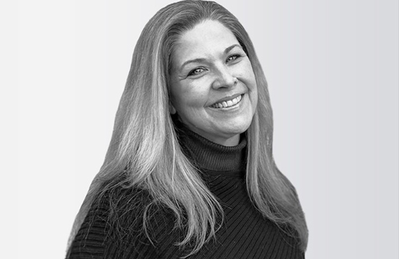 A smiling woman with long, light-colored hair wearing a dark turtleneck sweater.